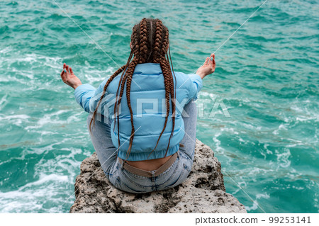 A woman in a blue jacket sits on a rock above a cliff above the sea, looking at the stormy ocean. Girl traveler rests, thinks, dreams, enjoys nature. Peace and calm landscape, windy weather. 99253141