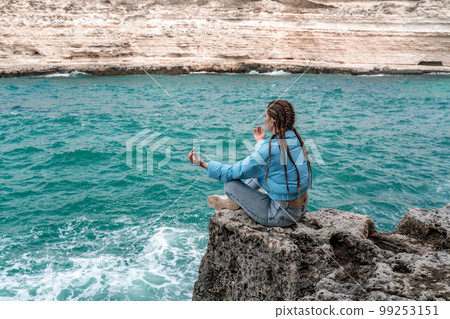 A woman in a blue jacket sits on a rock above a cliff above the sea, looking at the stormy ocean. Girl traveler rests, thinks, dreams, enjoys nature. Peace and calm landscape, windy weather. 99253151