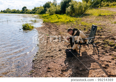Side view of bearded fisherman wearing raincoat preparing fishing gear on hook sitting on travel chair on river bank alone. Concept of lifestyle, leisure activity on nature. 99254369