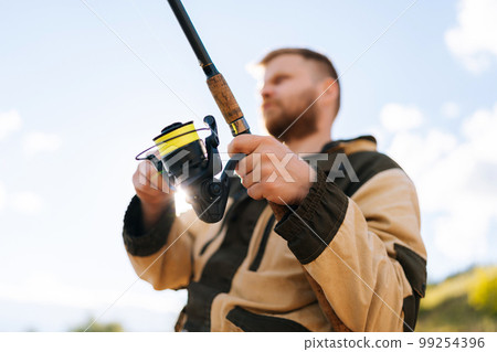 Shooting from below of fisherman holding casting rod wearing raincoat standing on bank waiting for bites on water river at summer day. Concept of lifestyle, leisure activity on nature. 99254396