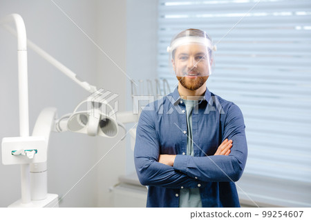 Medical safety. Portrait of young dentist doctor in face shield posing at workplace in modern clinic. Stomatolog looking at camera crossing his arms in front of him 99254607