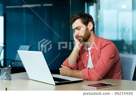Overtime, deadline. Tired young male programmer, freelancer sits in the office and tries to work at the laptop. Bored, he sits at the table and rests his head on his hand. 99255489