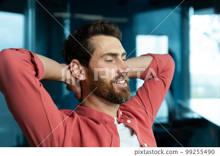A young male programmer, a freelancer, is resting from a hard day in the office at his desk. He sits on a chair, closes his eyes, puts his hands behind his head, meditates, dreams, smiles. 99255490