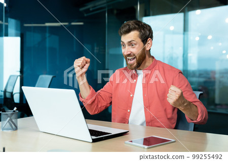 A young man sits in the office, looks at the laptop monitor and rejoices. Screams with happiness, makes victorious hand gestures. Received good news, successful deal, project, playing online games. A young man sits in the office, looks at the laptop monitor and rejoices. Screams with happiness, makes victorious hand gestures. Received good news, successful deal, project, playing online games. 99255492