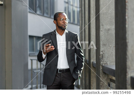 African American businessman in suit standing outside near office, bank, holding phone, hand in pocket. He is waiting for a business meeting, a client, a partner. 99255498