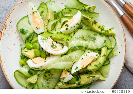 Healthy green avocado salad bowl with boiled eggs, sliced cucumbers, edamame beans, olive oil and herbs on ceramic plate top view, grey stone rustic table background Healthy green avocado salad bowl with boiled eggs, sliced cucumbers, edamame beans, olive oil and herbs on ceramic plate top view, grey stone rustic table background 99258119