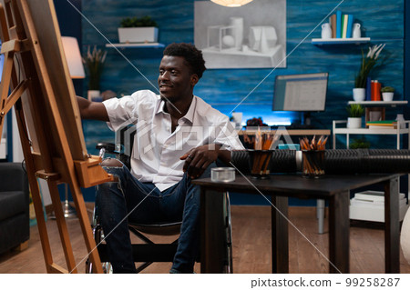 Smiling african american male cartoonist using colored pencils to sketch on easel at art studio. Wheelchair user drawing happily on canvas in bedroom. Person with artistic skills working in atelier. Smiling african american male cartoonist using colored pencils to sketch on easel at art studio. Wheelchair user drawing happily on canvas in bedroom. Person with artistic skills working in atelier. 99258287