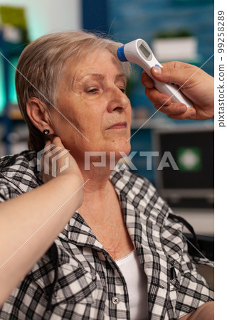 Senior woman in nursing home sitting while health care assistant takes temperature with infrared thermometer on forehead. Specialized healthcare for retired elderly people. Senior woman in nursing home sitting while health care assistant takes temperature with infrared thermometer on forehead. Specialized healthcare for retired elderly people. 99258289