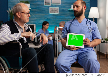 Medical nurse holding a green screen tablet with during a medical checkup of a nursing home resident patient. Elderly man in wheelchair talking to healthcare specialist in specialized center. 99258293