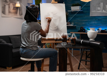 African american female professional teacher of drawing arts creating pencil sketch masterpiece of vase on canvas easel, sitting in home sketching atelier with creative tools at side. 99258310