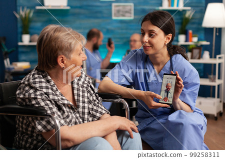 Senior woman in wheelchair having a medical consultation by video call with mobile phone. Female nurse in nursing home assisting patient during online doctor appointment. 99258311