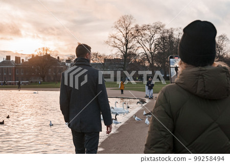 Rear view of woman wearing winter jacket photographing male friend with smartphone walking at lakeshore in park. Cloudy day during sunset in London 99258494