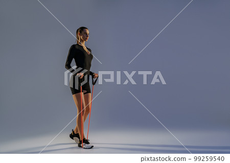 Strong woman in sportswear performs fitness exercises with resistance band on studio background 99259540