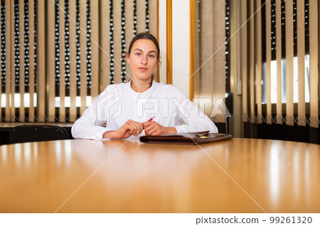 Portrait of a young woman sitting at a table in a conference room with a folder of documents Portrait of a young woman sitting at a table in a conference room with a folder of documents 99261320