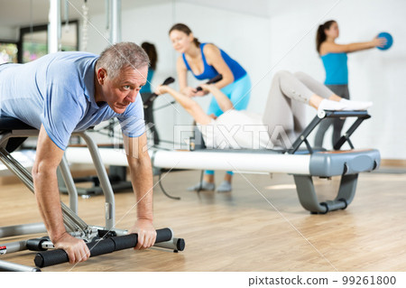 Concentrated old man in sportswear training on Pilates chair machine in rehabilitation center 99261800