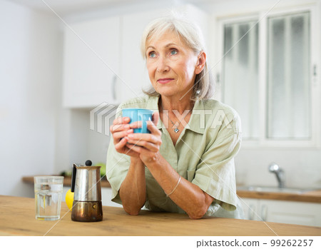 Positive old woman drinking coffee in kitchen at home Positive old woman drinking coffee in kitchen at home 99262257