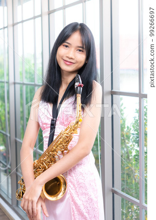 Young woman saxophonist with an instrument stands by the window. Cozy home interior. Concept of hobbies, development of talents,music school. Young woman saxophonist with an instrument stands by the window. Cozy home interior. Concept of hobbies, development of talents,music school. 99266697