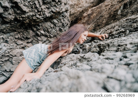 Sports Woman Climbing The Rock. Young woman With slim fit body climbing in volcanic basalt cave with beautiful sea view. The athlete girl trains in nature. Woman overcomes difficult climbing route. 99268969
