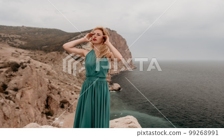 Redhead woman portrait. Curly redhead young caucasian woman with freckles looking at camera and smiling. Close up portrait cute woman in a mint long dress posing on a volcanic rock high above the sea 99268991
