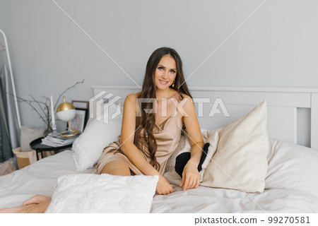 Positive sweet woman is sitting in bed leaning on pillows and smiling at the camera at home Positive sweet woman is sitting in bed leaning on pillows and smiling at the camera at home 99270581