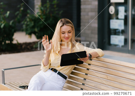 Stylish woman of thirty years old is sitting on a bench in the summer and talking on a tablet via video link 99270671
