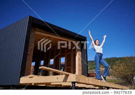 Happy woman on construction site standing on terrace at incomplete wooden frame house on sunny day with hands up. 99277639
