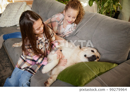 Two little cute happy girls, sisters sitting at home and playing with Golden retriever dog. Children look delighted and cheerful. Concept of friendship, family, care, animal Two little cute happy girls, sisters sitting at home and playing with Golden retriever dog. Children look delighted and cheerful. Concept of friendship, family, care, animal 99279964