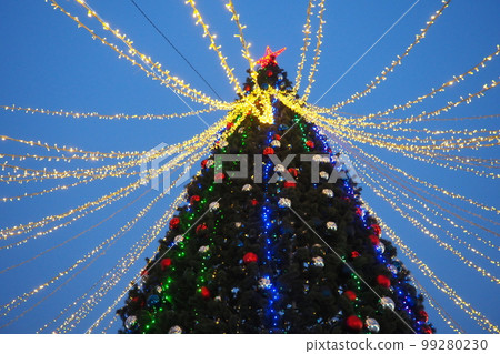 Multi-colored glass shiny balls on the New Year's city tree. Glowing garlands hang over the Christmas tree. Outdoor winter decoration. Festive design of the city square. Red, blue and yellow ball 99280230