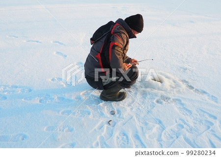 Fisherman on winter fishing. A man sits near a hole in a frozen lake waiting for a bite. Snow on ice. Bottom fishing for perch. Frosty January evening. Fisherman on winter fishing. A man sits near a hole in a frozen lake waiting for a bite. Snow on ice. Bottom fishing for perch. Frosty January evening. 99280234
