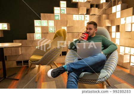 Caucasian bearded man sits in a comfortable chair drinks coffee and works on a laptop. Freelancer.  99280283