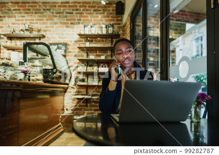 Portrait of a smiling young woman using laptop and talking on mobile phone in coffeeshop 99282787