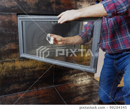 A man in a shirt and blue jeans cleans the heat-resistant glass of a modern fireplace. Close-up view of the hands. A man in a shirt and blue jeans cleans the heat-resistant glass of a modern fireplace. Close-up view of the hands. 99284345