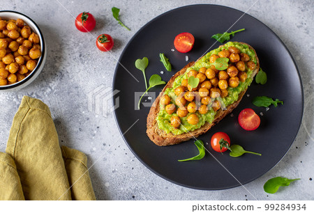 avocado toast with roasted chickpeas, herbs, cherry on a dark plate.  99284934