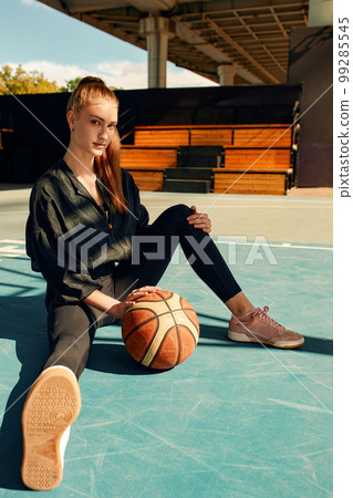 Girl basketball player smiles and sits on the sports field with a basketball and looks at the camera. Sport, fitness, active lifestyle. Girl basketball player smiles and sits on the sports field with a basketball and looks at the camera. Sport, fitness, active lifestyle. 99285545