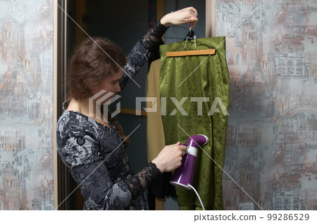 Woman processes clothes with a manual steamer against the backdrop of a home dressing room 99286529