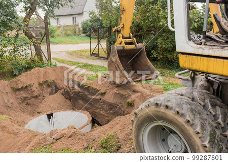 A bucket of excavator with a pile of sand and earth buries sewer concrete rings in the industrial zone 99287801