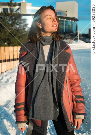 a young beautiful girl in a sheepskin coat on an ice rink against the backdrop of the metropolis and the rays of the sun 99288859