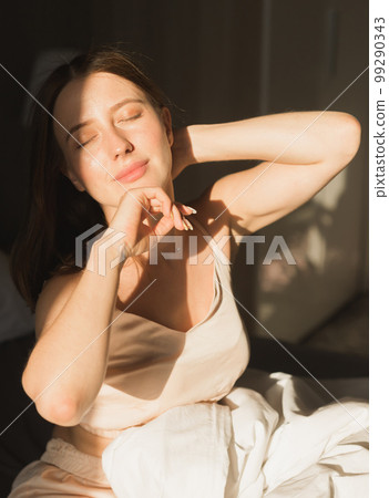 Girl is waking up in morning stretches in bed and sun shines from the window. Happy young woman greets new sunny day 99290343