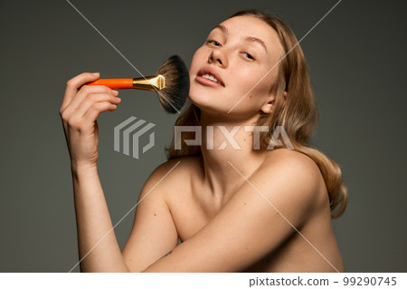 Half-length portrait of young beautiful girl with bare shoulders powdering with makeup brush. Concept of beauty, natural make-up, skin care, healthy look 99290745