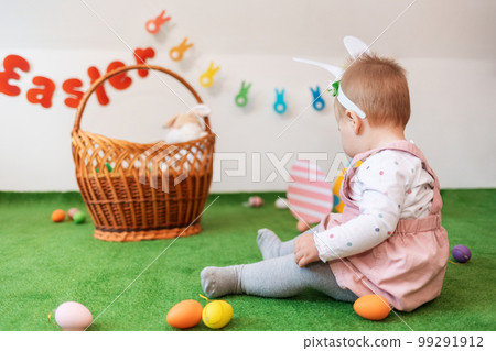A baby girl with rabbit ears, sitting on an artificial lawn looking at the background with a basket and eggs. Rear view. Easter holiday concept 99291912