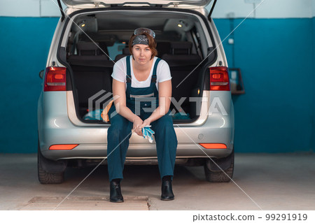 A female mechanic in coveralls is sitting in the open trunk of a car. Work at garage 99291919