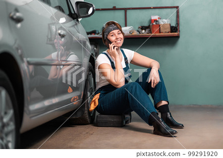 Happy young woman mechanic is sitting leaning on a car in an auto repair shop and calling at cellphone. The concept of women's work in male professions and garage work 99291920