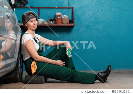 Pretty young woman mechanic is sitting leaning on a car in an auto repair shop. The concept of women's work in male professions Pretty young woman mechanic is sitting leaning on a car in an auto repair shop. The concept of women's work in male professions 99291921
