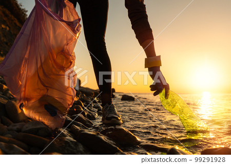Pollution of ocean. A volunteer take a plastic bottle by the sea or river holding a plastic trash bag. Coastal cleanup and garbage collection for recycling. World environment day and Earth day concept 99291928