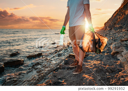 Close up of volunteer stand at the wild beach with a plastic bag and a bottle in his hands. In the background, the sea and the sunset. Copy space. The concept of cleaning the beach from pollution Close up of volunteer stand at the wild beach with a plastic bag and a bottle in his hands. In the background, the sea and the sunset. Copy space. The concept of cleaning the beach from pollution 99291932