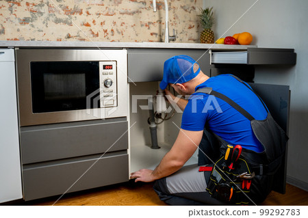 Uniformed plumber is using a wrench to repair a water pipe under the sink. Maintenance concept: eliminate leaks in the water supply, clean clogged pipes from dirt Uniformed plumber is using a wrench to repair a water pipe under the sink. Maintenance concept: eliminate leaks in the water supply, clean clogged pipes from dirt 99292783