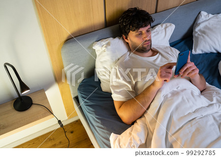 Top view of a black-haired man who uses a smartphone late at night, turns off the light and goes to bed 99292785