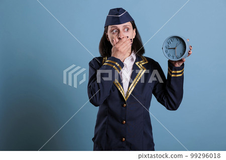 Anxious stewardess holding retro alarm clock, running late at airport, front view studio medium shot. Confused flight attendant overslept, time management, deadline, schedule concept Anxious stewardess holding retro alarm clock, running late at airport, front view studio medium shot. Confused flight attendant overslept, time management, deadline, schedule concept 99296018