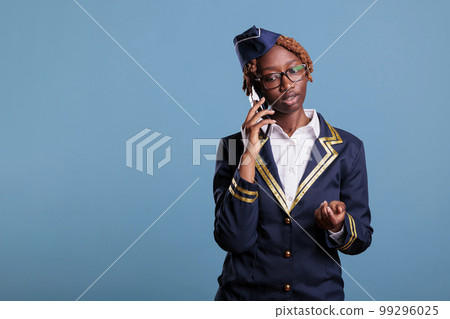 Concentrated female flight attendant talking on the phone dressed in uniform and eyeglasses. Stewardess looks serious while using mobile device in studio shot against blue background. 99296025