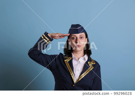 Portrait of flight attendant doing aviation honour salut in front of camera. Serious stewardess wearing professional flight uniform standing in studio with blue background, medium shot 99296028